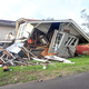 A man wearing red shorts holds his head in distress, standing in front of his collapsed house